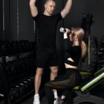 Fitness instructor conducts personal training for a girl with dumbbell in front of the mirror